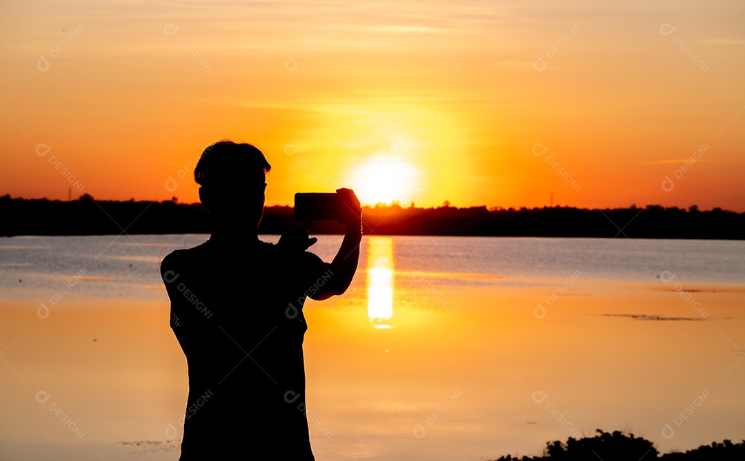 Silhueta, homem usando smartphone tira foto do pôr do sol no lago