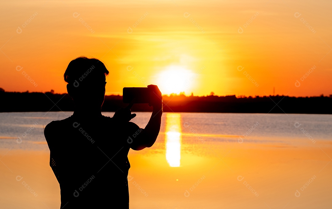 Silhueta, homem usando smartphone tira foto do pôr do sol no lago