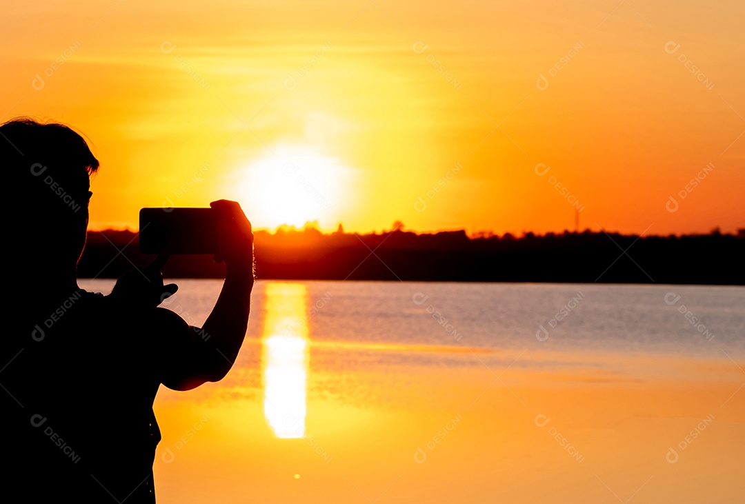 Silhueta, homem usando smartphone tira foto do pôr do sol no lago