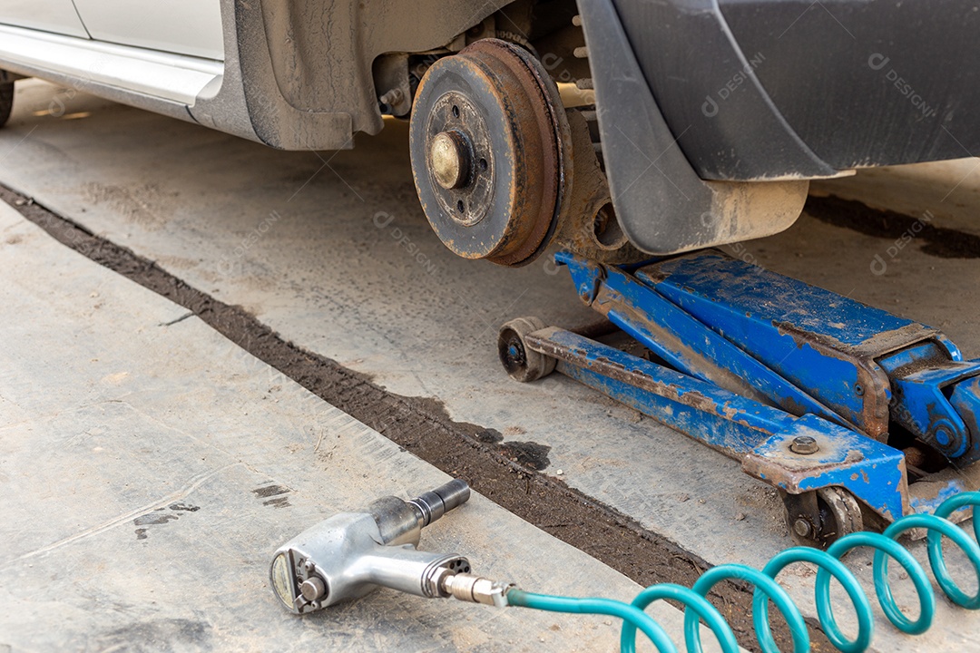 Carro sem roda levantado com macaco hidráulico e chave de impacto pneumática em uma oficina mecânica ao ar livre