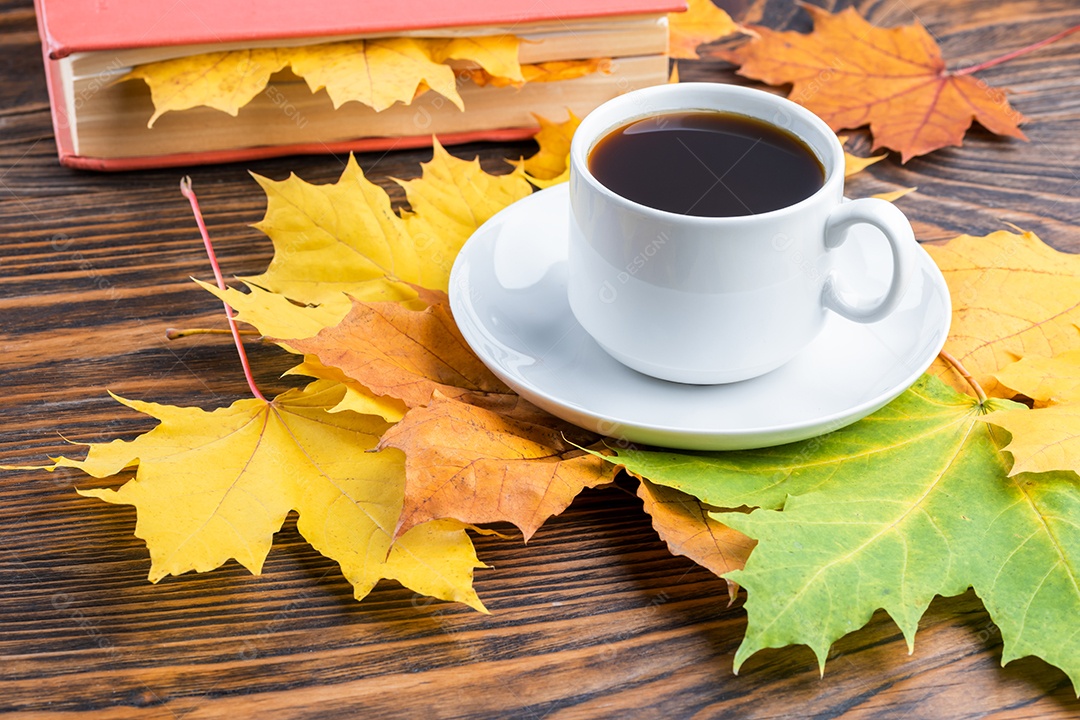 Xícara de café sobre mesa de madeira com livro e folhas coloridas de bordo outonais