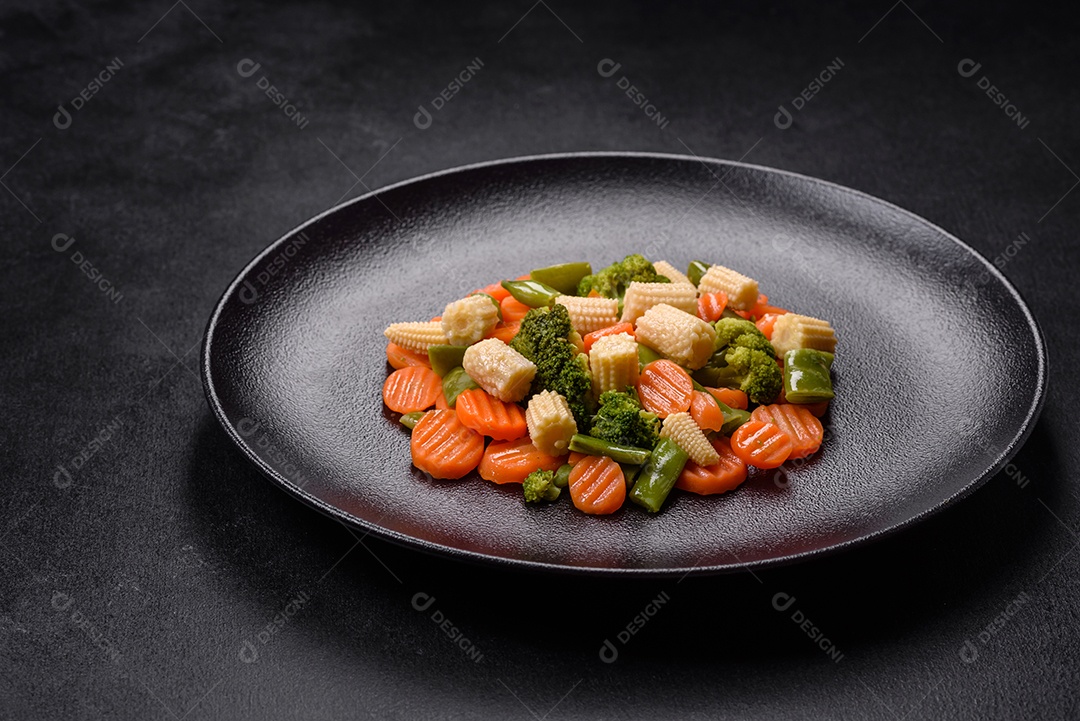 Uma mistura de vegetais: cenouras, pequenas espigas de milho, grãos de aspargos cozidos no vapor em um prato de cerâmica preta