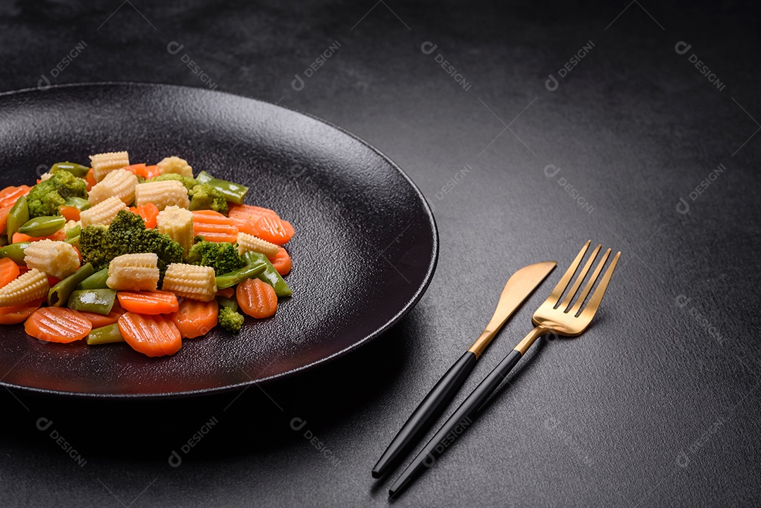 Uma mistura de vegetais: cenouras, pequenas espigas de milho, grãos de aspargos cozidos no vapor em um prato de cerâmica preta