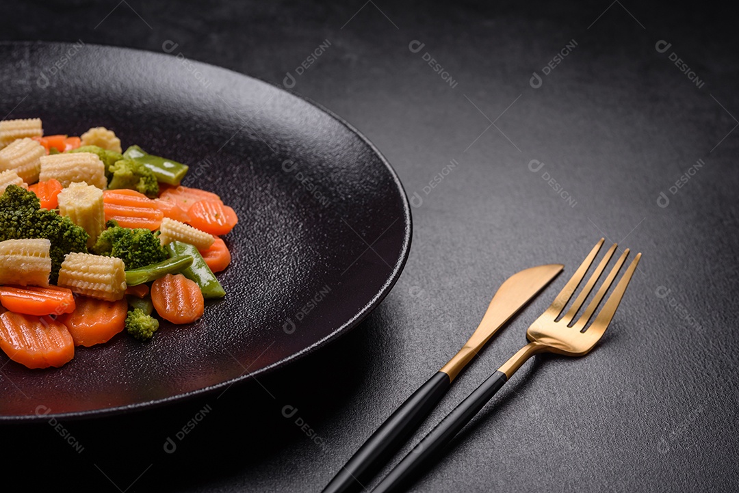 Uma mistura de vegetais: cenouras, pequenas espigas de milho, grãos de aspargos cozidos no vapor em um prato de cerâmica preta