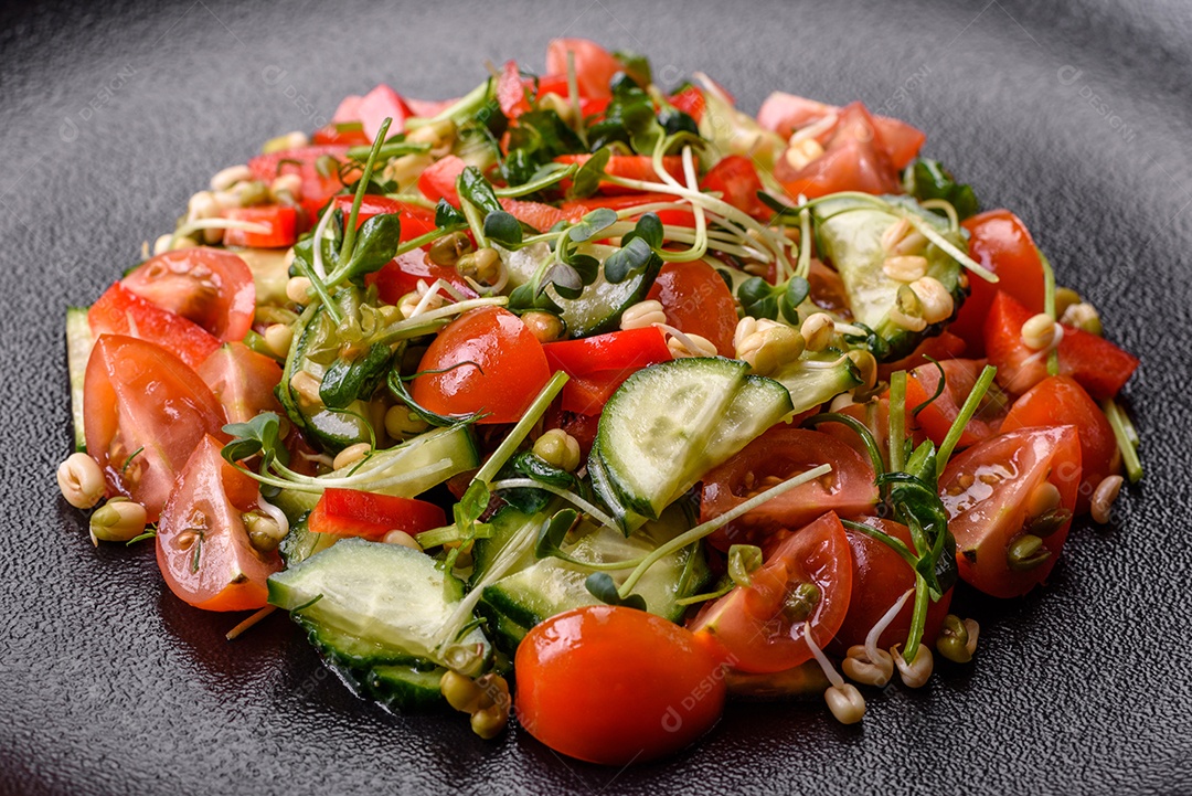 Deliciosa salada fresca e suculenta de tomate, pimentão, pepino, microgreens e feijão-mungo germinado em uma placa de cerâmica preta sobre um fundo escuro de concreto