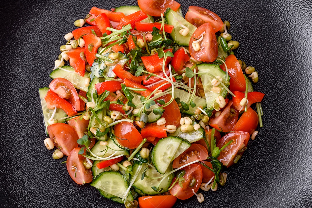 Deliciosa salada fresca e suculenta de tomate, pimentão, pepino, microgreens e feijão-mungo germinado em uma placa de cerâmica preta sobre um fundo escuro de concreto