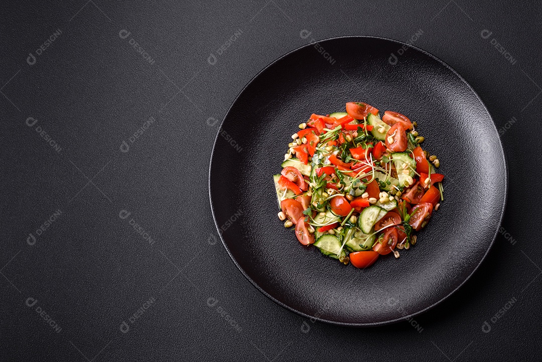 Deliciosa salada fresca e suculenta de tomate, pimentão, pepino, microgreens e feijão-mungo germinado em uma placa de cerâmica preta sobre um fundo escuro de concreto