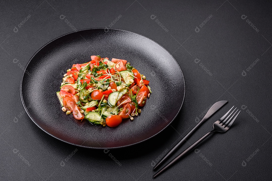 Deliciosa salada fresca e suculenta de tomate, pimentão, pepino, microgreens e feijão-mungo germinado em uma placa de cerâmica preta sobre um fundo escuro de concreto