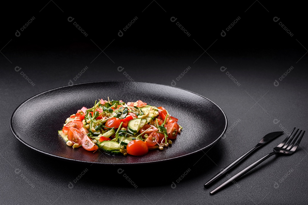 Deliciosa salada fresca e suculenta de tomate, pimentão, pepino, microgreens e feijão-mungo germinado em uma placa de cerâmica preta sobre um fundo escuro de concreto
