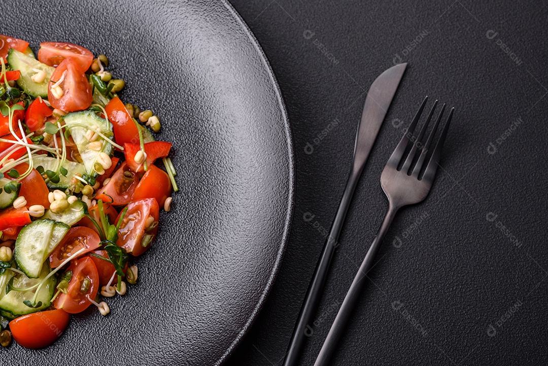 Deliciosa salada fresca e suculenta de tomate, pimentão, pepino, microgreens e feijão-mungo germinado em uma placa de cerâmica preta sobre um fundo escuro de concreto