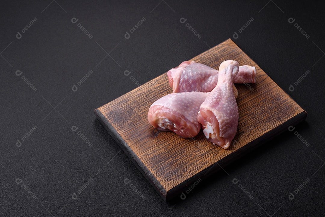 Três pernas de frango cru com especiarias e ervas em uma tábua de madeira contra um fundo escuro de concreto
