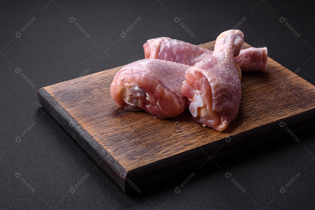Três pernas de frango cru com especiarias e ervas em uma tábua de madeira contra um fundo escuro de concreto