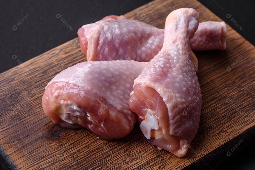 Três pernas de frango cru com especiarias e ervas em uma tábua de madeira contra um fundo escuro de concreto