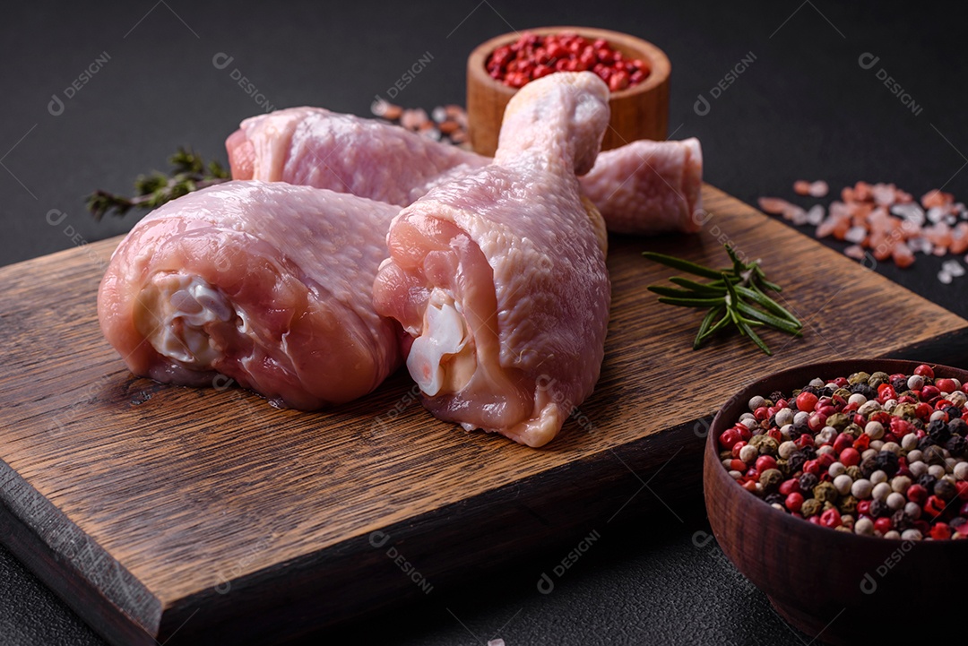 Três pernas de frango cru com especiarias e ervas em uma tábua de madeira contra um fundo escuro de concreto