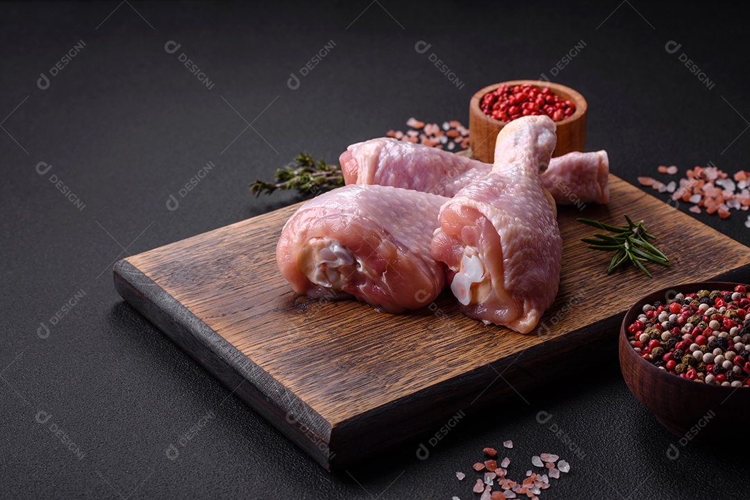 Três pernas de frango cru com especiarias e ervas em uma tábua de madeira contra um fundo escuro de concreto
