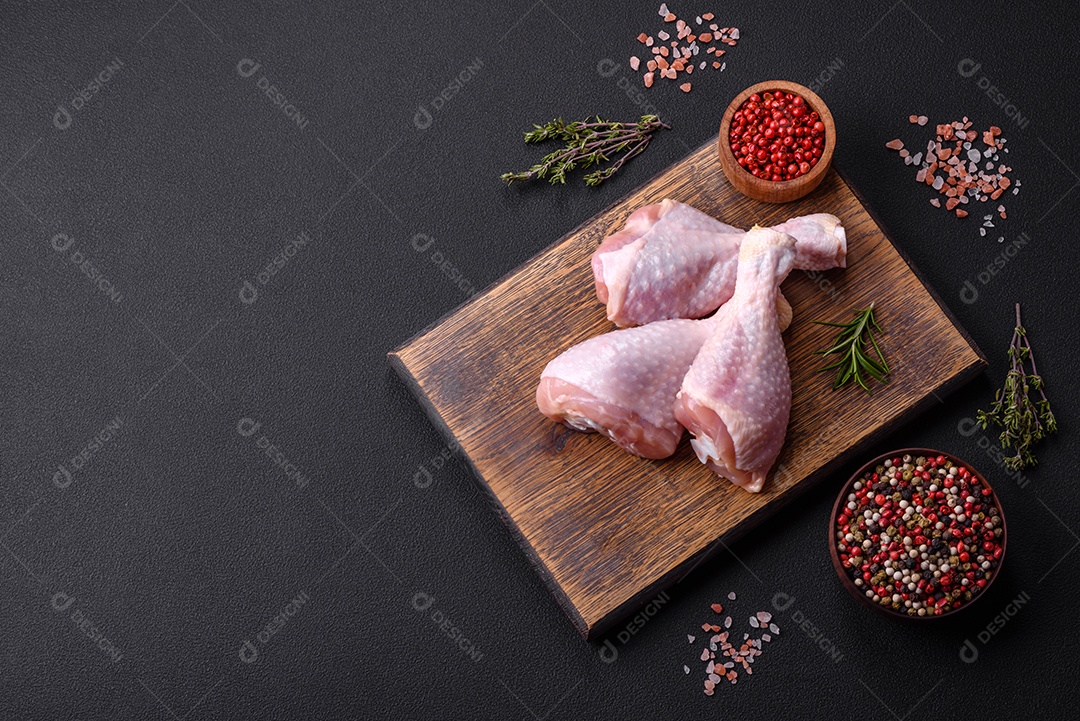 Três pernas de frango cru com especiarias e ervas em uma tábua de madeira contra um fundo escuro de concreto