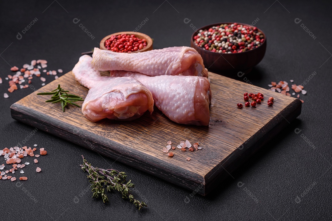 Três pernas de frango cru com especiarias e ervas em uma tábua de madeira contra um fundo escuro de concreto
