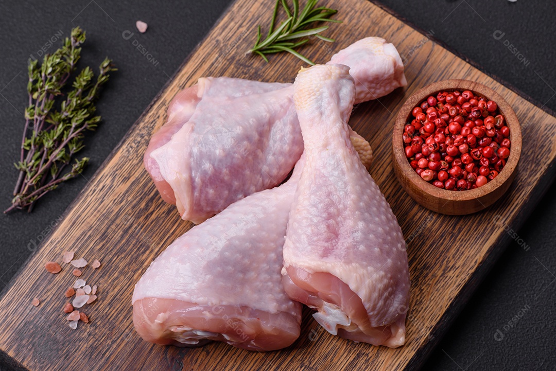 Três pernas de frango cru com especiarias e ervas em uma tábua de madeira contra um fundo escuro de concreto