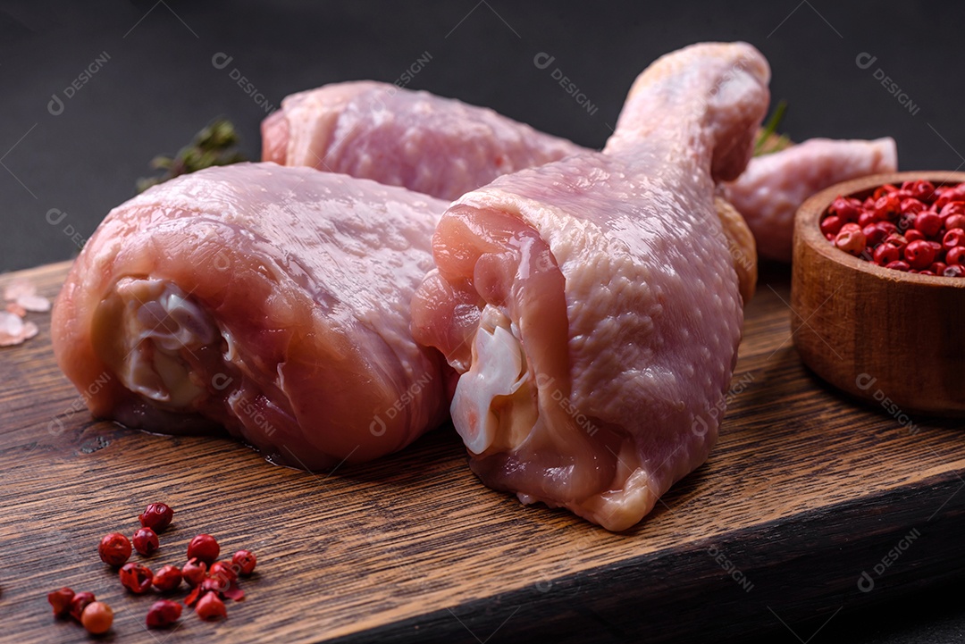 Três pernas de frango cru com especiarias e ervas em uma tábua de madeira contra um fundo escuro de concreto