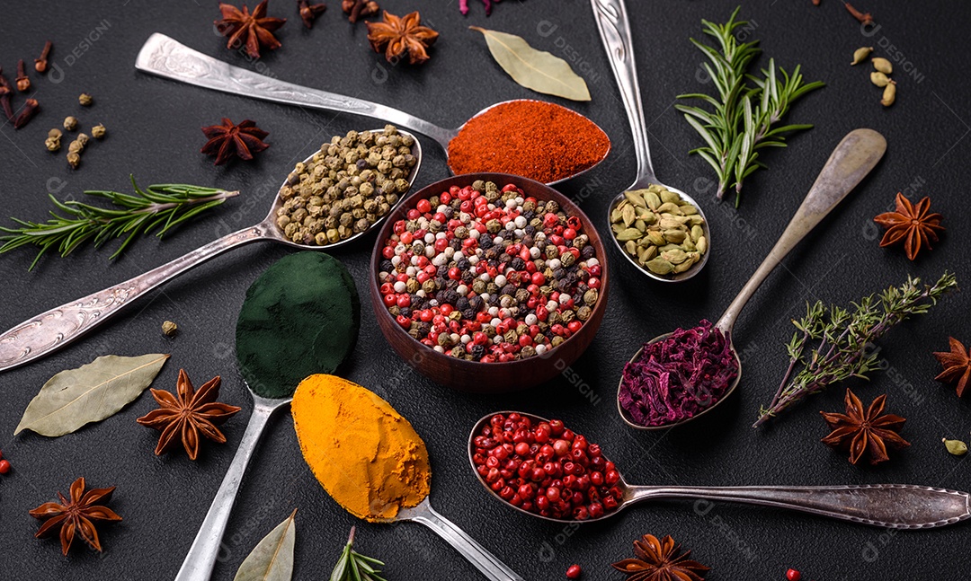 Variação de especiarias em colheres de metal páprica, açafrão, cardamomo, uma mistura de pimenta da Jamaica, tomilho, alecrim e sal sobre um fundo escuro de concreto