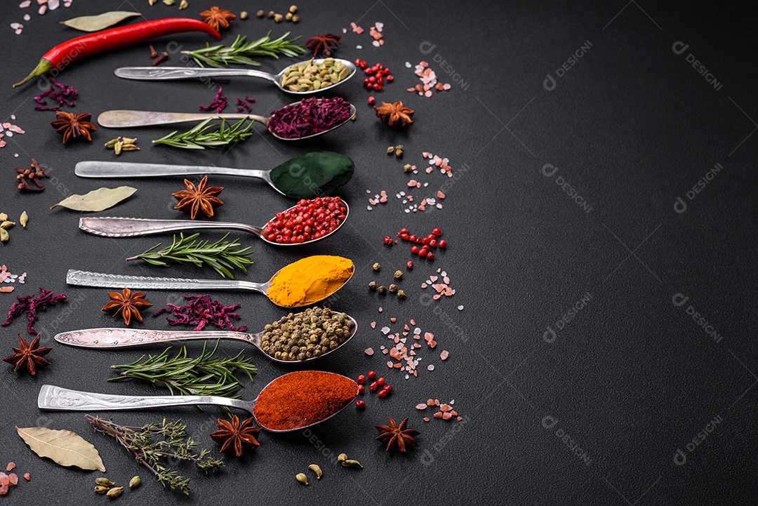 Variação de especiarias em colheres de metal páprica, açafrão, cardamomo, uma mistura de pimenta da Jamaica, tomilho, alecrim e sal sobre um fundo escuro de concreto
