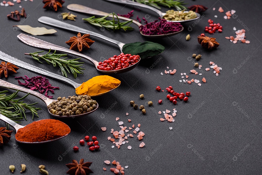 Variação de especiarias em colheres de metal páprica, açafrão, cardamomo, uma mistura de pimenta da Jamaica, tomilho, alecrim e sal sobre um fundo escuro de concreto