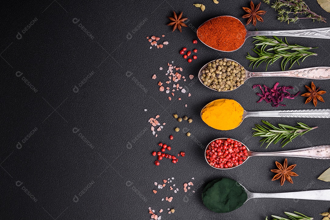 Variação de especiarias em colheres de metal páprica, açafrão, cardamomo, uma mistura de pimenta da Jamaica, tomilho, alecrim e sal sobre um fundo escuro de concreto