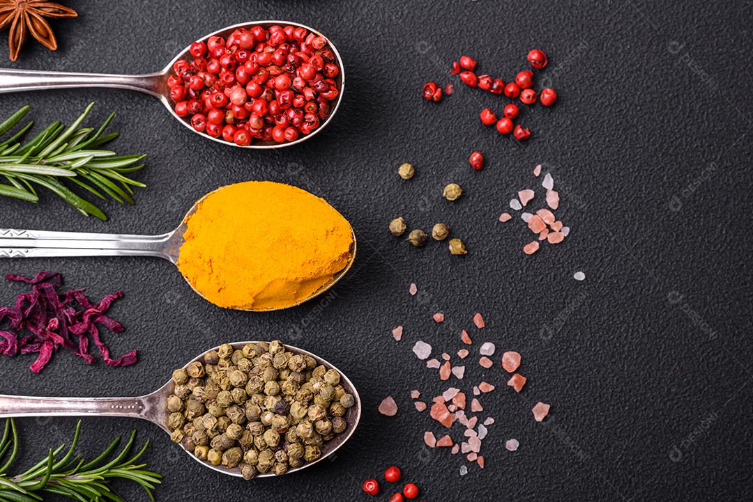 Variação de especiarias em colheres de metal páprica, açafrão, cardamomo, uma mistura de pimenta da Jamaica, tomilho, alecrim e sal sobre um fundo escuro de concreto
