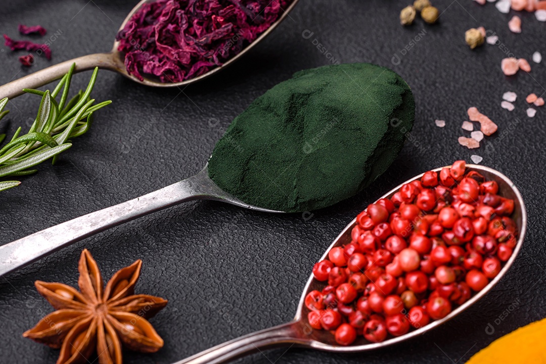 Variação de especiarias em colheres de metal páprica, açafrão, cardamomo, uma mistura de pimenta da Jamaica, tomilho, alecrim e sal sobre um fundo escuro de concreto