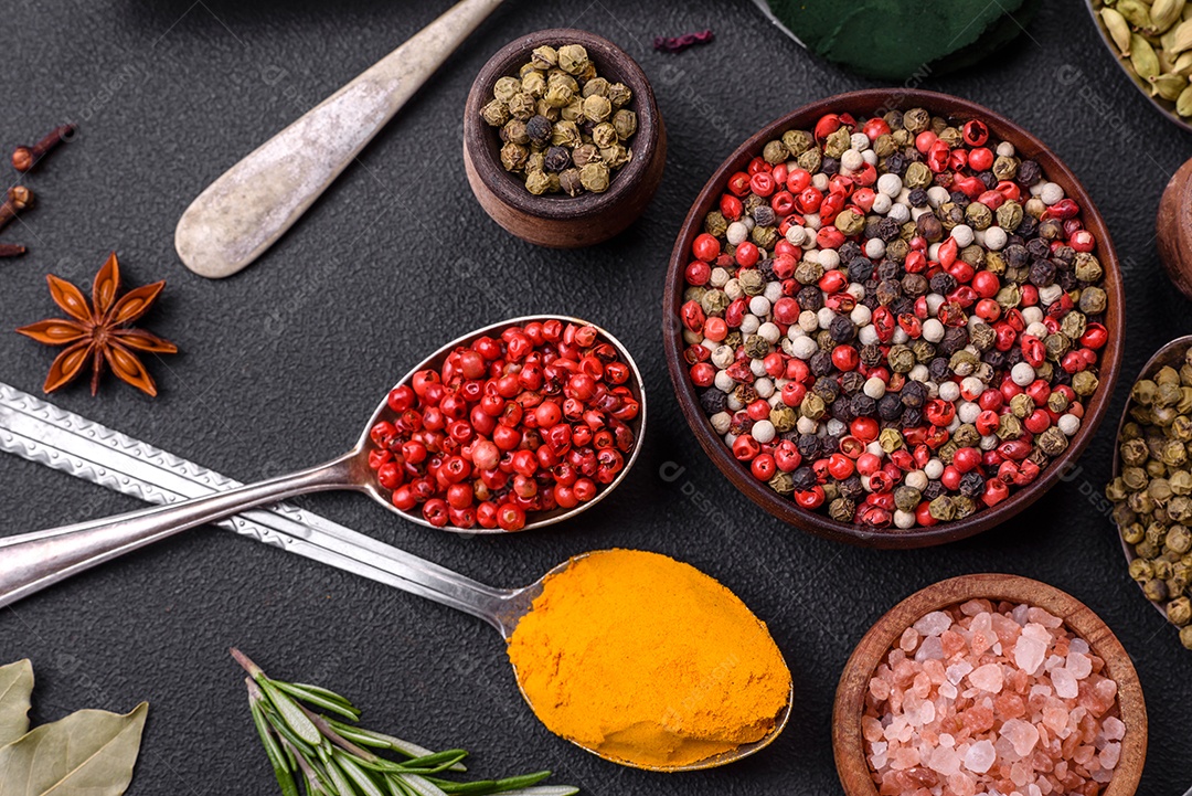 Variação de especiarias em colheres de metal páprica, açafrão, cardamomo, uma mistura de pimenta da Jamaica, tomilho, alecrim e sal sobre um fundo escuro de concreto