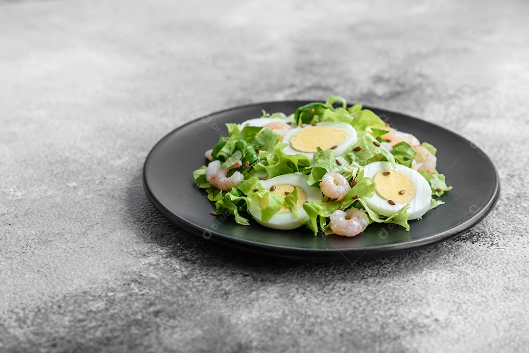 Deliciosa salada fresca e saudável com camarão, ovo, alface e sementes de linho em uma placa de cerâmica preta sobre um fundo de concreto
