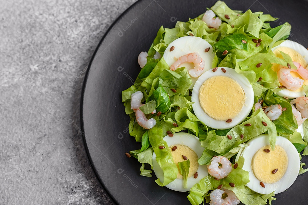 Deliciosa salada fresca e saudável com camarão, ovo, alface e sementes de linho em uma placa de cerâmica preta sobre um fundo de concreto