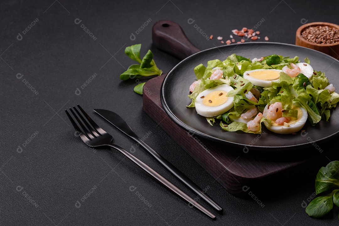 Deliciosa salada fresca e saudável com camarão, ovo, alface e sementes de linho em uma placa de cerâmica preta sobre um fundo de concreto