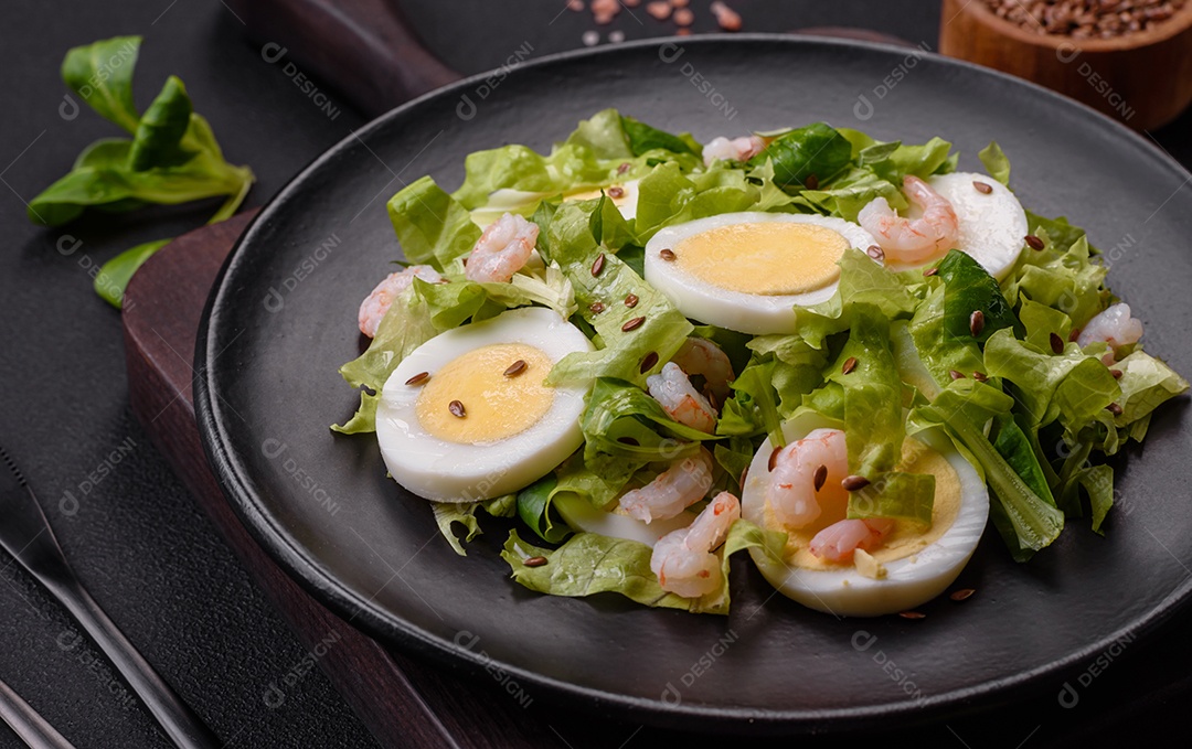 Deliciosa salada fresca e saudável com camarão, ovo, alface e sementes de linho em uma placa de cerâmica preta sobre um fundo de concreto