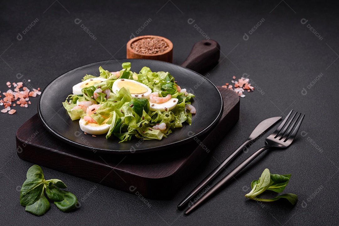 Deliciosa salada fresca e saudável com camarão, ovo, alface e sementes de linho em uma placa de cerâmica preta sobre um fundo de concreto