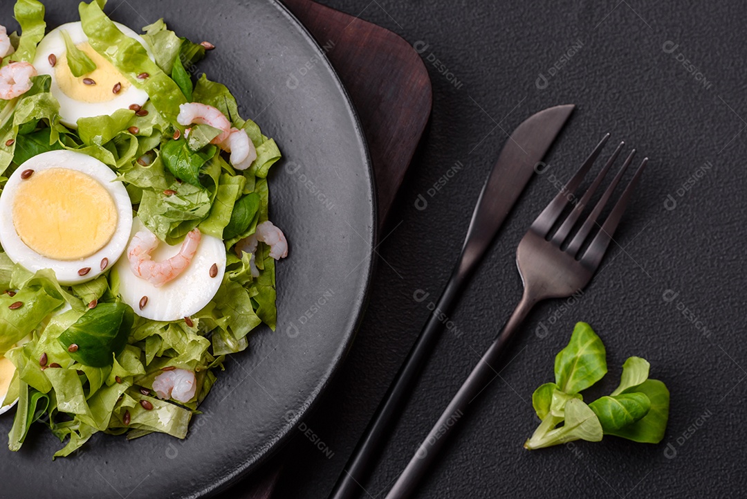 Deliciosa salada fresca e saudável com camarão, ovo, alface e sementes de linho em uma placa de cerâmica preta sobre um fundo de concreto