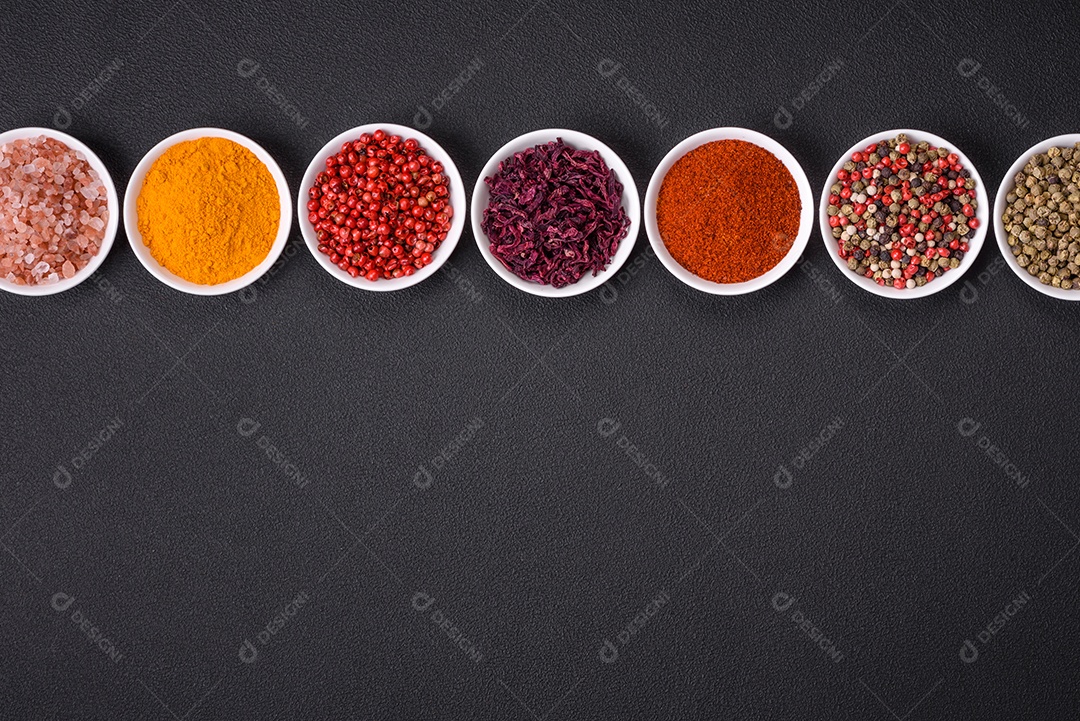 Composição composta por variações de especiarias em tigelas brancas e colheres de metal sobre fundo escuro de concreto