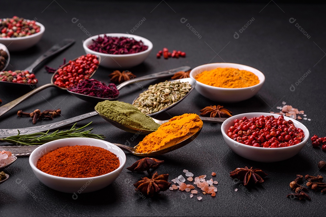 Composição composta por variações de especiarias em tigelas brancas e colheres de metal sobre fundo escuro de concreto