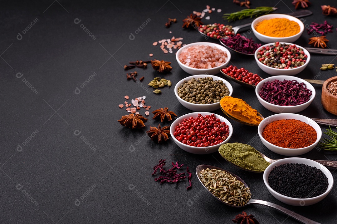 Composição composta por variações de especiarias em tigelas brancas e colheres de metal sobre fundo escuro de concreto