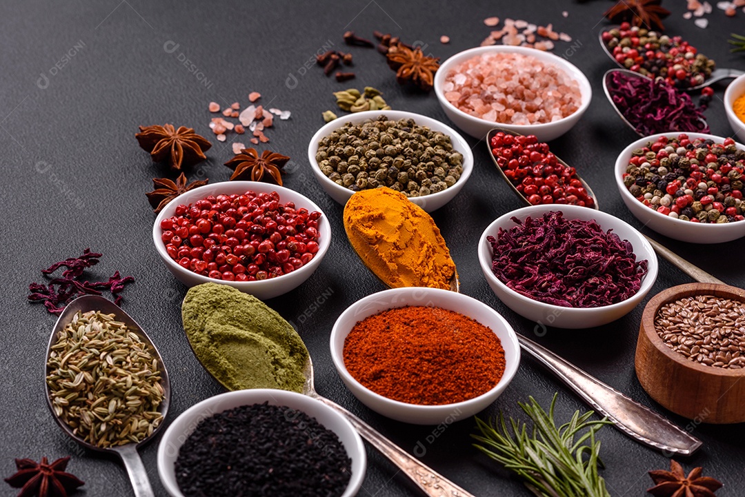 Composição composta por variações de especiarias em tigelas brancas e colheres de metal sobre fundo escuro de concreto