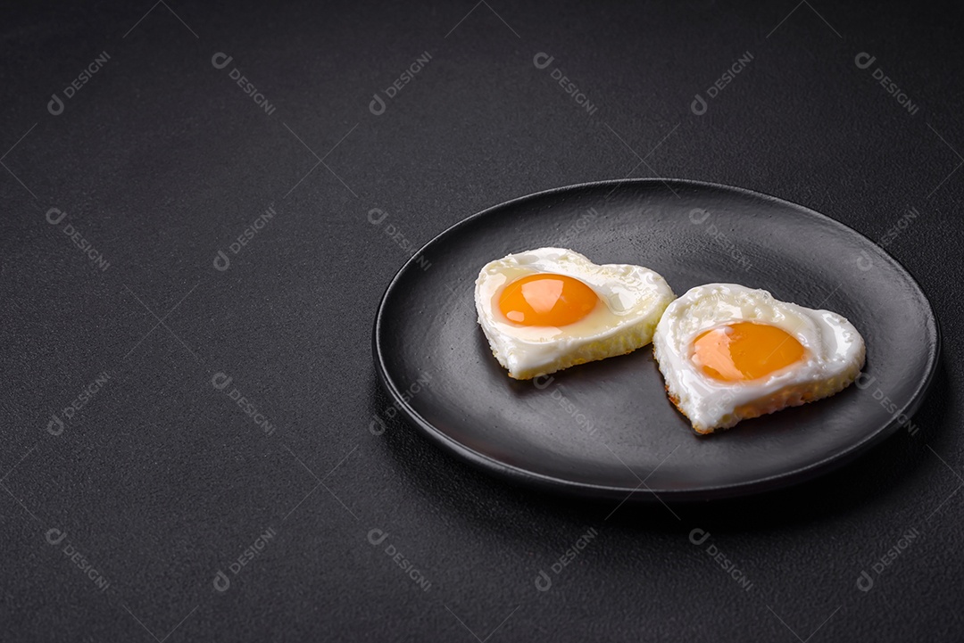 Dois ovos fritos em forma de coração em uma placa de cerâmica preta sobre um fundo escuro de concreto. Café da manhã para o dia dos namorados