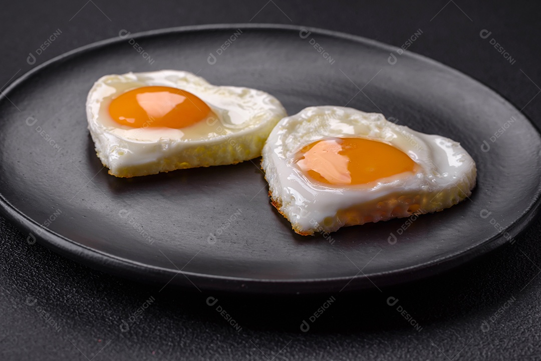 Dois ovos fritos em forma de coração em uma placa de cerâmica preta sobre um fundo escuro de concreto. Café da manhã para o dia dos namorados