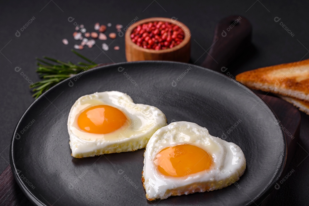 Dois ovos fritos em forma de coração em uma placa de cerâmica preta sobre um fundo escuro de concreto. Café da manhã para o dia dos namorados