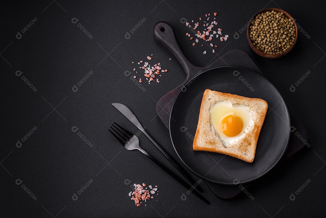 Ovo frito em forma de coração em torradas de pão com sementes de gergelim, sementes de linho e sementes de abóbora em uma placa preta sobre um fundo escuro de concreto