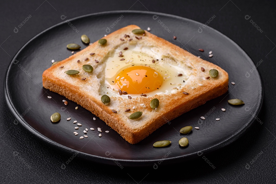 Ovo frito em forma de coração em torradas de pão com sementes de gergelim, sementes de linho e sementes de abóbora em uma placa preta sobre um fundo escuro de concreto