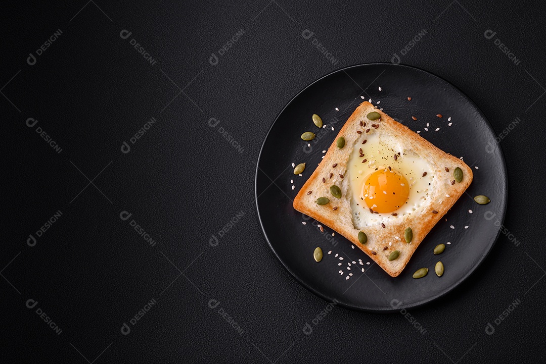 Ovo frito em forma de coração em torradas de pão com sementes de gergelim, sementes de linho e sementes de abóbora em uma placa preta sobre um fundo escuro de concreto