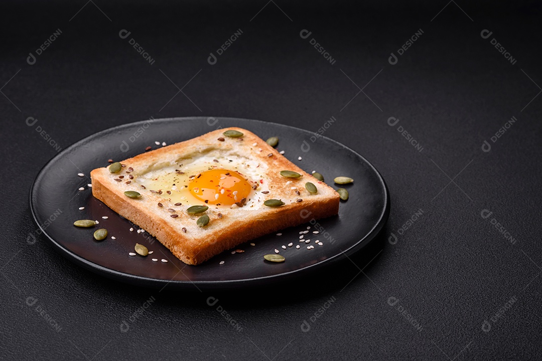 Ovo frito em forma de coração em torradas de pão com sementes de gergelim, sementes de linho e sementes de abóbora em uma placa preta sobre um fundo escuro de concreto