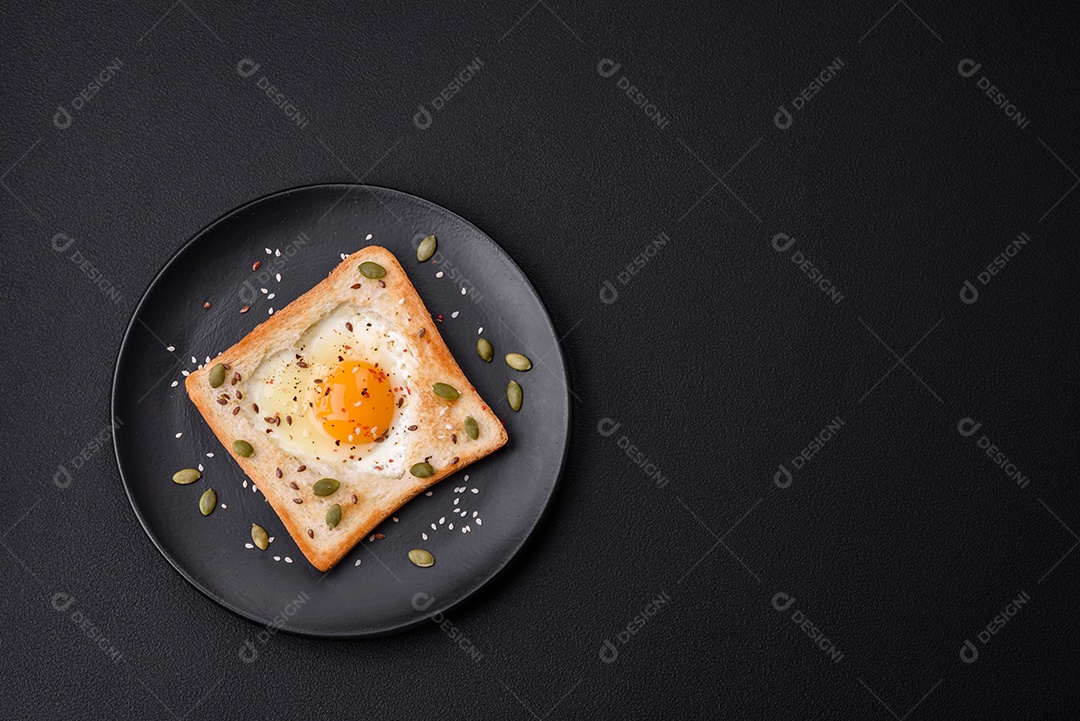 Ovo frito em forma de coração em torradas de pão com sementes de gergelim, sementes de linho e sementes de abóbora em uma placa preta sobre um fundo escuro de concreto