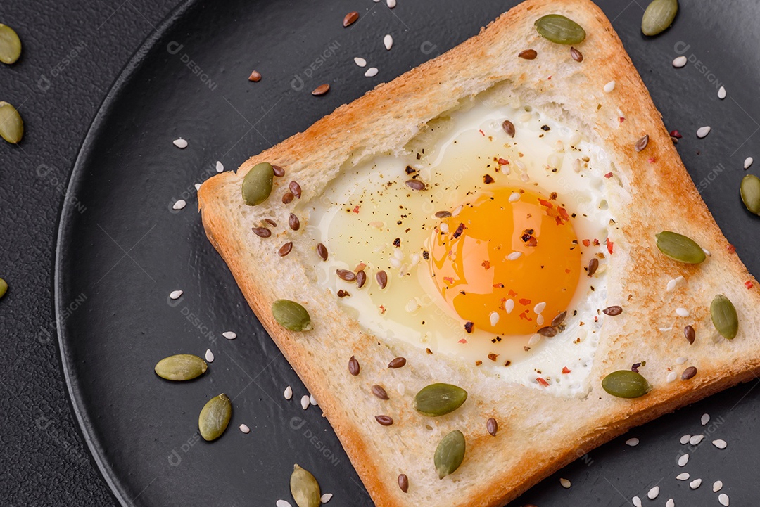Ovo frito em forma de coração em torradas de pão com sementes de gergelim, sementes de linho e sementes de abóbora em uma placa preta sobre um fundo escuro de concreto