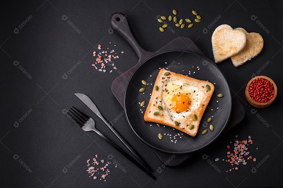Ovo frito em forma de coração em torradas de pão com sementes de gergelim, sementes de linho e sementes de abóbora em uma placa preta sobre um fundo escuro de concreto